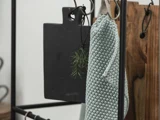 Et sort skærebræt, en grøn urtekvist og et lysegrønt køkkenhåndklæde med struktur hænger på kroge fra et sort metalstativ.