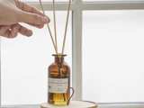 En hånd placerer en sivpind i en diffusorflaske af brunt glas med mærket ALTUM Meadow Honey & Lemon, som står på en træplade ved et lyst vindue.