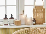En hyggelig vindueskarm med håndsæbe, lotion, stearinlys, en flettet lanterne, små beholdere og en blomstret pude på en kurvestol.