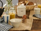 Et morgenbord med et blødkogt æg, en stribet skål, appelsinjuice, croissanter, en krukke med påskriften Sweets og blåternede servietter på et rustikt træbord.