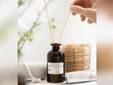 En hånd placerer siv i en brun ALTUM-diffusorflaske mærket Meadow, Honey & Lemon med et stearinlys, et rør og en sten i nærheden på en lys overflade.