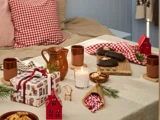 En hyggelig julescene med en seng, et festligt dækket bord med krus, småkager, stearinlys, indpakkede gaver, fyrretræsgrene og rødternede detaljer.