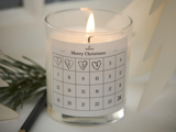 Et tændt stearinlys i en glasholder med en adventskalenderetiket med titlen Merry Christmas, der viser de nummererede dage 1 til 24. Fyrretræ og en blyant ligger i nærheden.