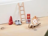 Miniaturejulescene med en lille rød nissehue, grå sko, en træstige, stablede gaveæsker, en lanterne og en trækælk på et lyst trægulv.