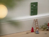 En lille grøn dør er fastgjort til en hvid væg over en fodliste. Nedenfor er der en lille træstige, et rødt miniaturetræ og små grå sko på et trægulv.