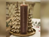 Et brunt adventslys med nummererede dage tændes og placeres på en guldbakke, flankeret af to dekorative guldjuletræsornamenter.
