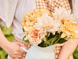 En person i gingham-kjole holder en hvid kande fyldt med cremefarvede og gule blomster, bl.a. hortensiaer og georginer.