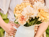 En person i gingham-kjole holder en hvid kande fyldt med cremefarvede og gule blomster med grønne blade.