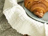 Et nærbillede af en struktureret hvid klud, der er draperet over en overflade ved siden af en tallerken med en croissant på.