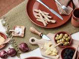 En rustik borddækning med brødstænger, oliven, ananas, brombær, rødløg, salt, en kniv og brune tallerkener på en grøn vævet dækkeserviet.