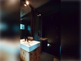 Et moderne badeværelse med træbordplade, hvid håndvask, stort spejl og varm ovenlys; et toilet og en anden håndvask er svagt synlige i baggrunden.