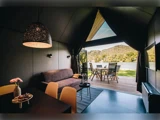 Moderne glampingtelt med sofa, spisebord og pendellampe, der åbner op til en terrasse med udendørs siddepladser med udsigt over en sø og skovklædte bakker.