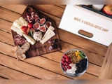 Et træbræt med ost, kødskiver, brød, figner, tomater og dip ved siden af en skål granola med frugt på et træbord. På en kasse står der "HAVE A CHOCOLASTIC DAY.".