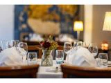 Elegant restaurantbord dækket med vinglas, vandglas, hvide servietter og en vase med røde blomster, med blød belysning og sløret baggrund.