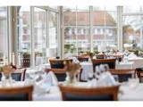 Lys, elegant restaurant med store vinduer, opdækkede borde med hvide duge, glas og blomsterdekorationer med udsigt over en gade udenfor.