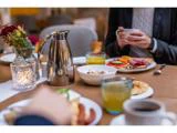 Et bord er dækket til morgenmad med kaffe, juice, müsli, brød, pålæg og frugt. To personer sidder, den ene holder et krus. En vase med blomster og et stearinlys står også på bordet.