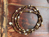 En lang perlekæde med brune og hvide perler er rullet op på en rustik træoverflade. Teksten Lang unika halskæde står i nederste højre hjørne.