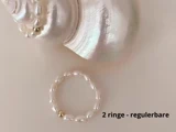 En fin perlering med en guldperle vises på en cremefarvet overflade ved siden af en hvid muslingeskal. Teksten 2 ringe - regulerbare står i nederste højre hjørne.