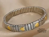 Et sølvfarvet justerbart armbånd i rustfrit stål med guldbelagte hjertecharms på en beige stofbaggrund. Den danske tekst lyder: Regulerbart stålarmbånd med forgyldte hjerter.