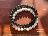 To perlearmbånd af ædelsten - et med sorte perler og et med hvide, marmorerede perler - på en træoverflade med sollys og skygge. Tekst: 2 armbånd i ædelsten.