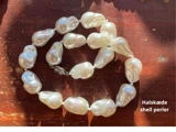 En halskæde af store, uregelmæssigt formede hvide skalperler vises på en træoverflade med sollys og skygger. Teksten lyder: Halskæde shell perler.