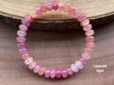 Et perlearmbånd af facetterede rosa og lys ferskenfarvede agatsten ligger på en træplade. Teksten Lyserød Agat står i nederste højre hjørne.
