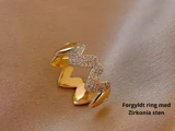 En guldbelagt ring med zigzag-design, prydet med zirkonia-sten, placeret på et beige stof. Teksten lyder: Forgyldt ring med Zirkonia sten.