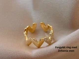 Guldbelagt ring med hjerteformede mønstre, det ene besat med små zirkonsten, placeret på et blødt beige stof. Tekst: Forgyldt ring med Zirkonia sten.