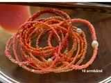 Et bundt med 10 perlearmbånd i nuancer af pink, orange og guld, nogle med små perlelignende perler, vist på en skinnende metallisk overflade. Tekst: 10 armbånd.