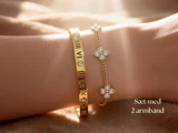 Et håndled med to guldarmbånd: et med romertal og et med små blomsterformede charms med krystaller. Teksten lyder: Sæt med 2 armbånd.