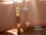 Nærbillede af et håndled, der bærer to guldarmbånd: et med romertal og krystaller og et med blomsterformede charms. Teksten siger Sæt med 2 armbånd.