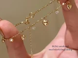 En hånd holder et fint guldkædearmbånd prydet med små hjertecharms og funklende ædelstene. Den danske tekst på billedet lyder: BLING armbånd med stærk forgyldning.