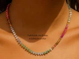 En kvinde bærer en delikat halskæde lavet af små, flerfarvede ædelstensperler. Teksten på billedet lyder: Halskæde med små ædelstens perler.