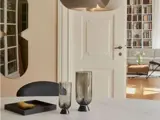 En moderne spiseplads med et rundt bord, to røgfarvede glasvaser, en bakke med en pære og et kig ind i en hyggelig stue med bogreoler og en sofa.