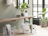 Et minimalistisk badeværelse med potteplanter, hvide trådkurve, foldede håndklæder, rengøringsflasker og en hvid spand på en træbænk nær store vinduer.