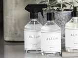 Tre ALTUM-rengøringsflasker med hvide etiketter står på en disk ved siden af en grå urtepotte med grønne blade. Den ene flaske har en spraydyse.