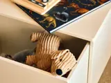 I en åben hvid skuffe ligger et ribbet, beige tøjdyr i form af en hund, og oven på skuffen ligger en bog med dinosaur-tema og et stykke trælegetøj.