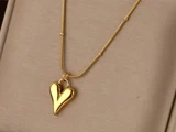 En guldhalskæde med et skinnende, asymmetrisk hjerteformet vedhæng på en beige stofbaggrund.