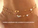 Nærbillede af en ankel iført en dobbelt guldankelkæde med hjerteformede charms og små perler. Den danske tekst nedenunder lyder: Dobbelt ankelkæde eller armbånd til bred håndled.