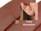 Nærbillede af en lille margueritformet ørering, der holdes i fingrene, med et indslag, der viser tre margueritøreringe båret på et øre. Teksten lyder Daisy Ørestikker.