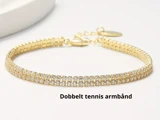 Et dobbeltradet tennisarmbånd i guld med funklende sten og en justerbar lås, vist på en lys overflade. Teksten lyder: Dobbelt tennis armbånd.