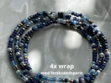 Fire tråde af blåt, sort og hvidt perlearmbånd med teksten "4x wrap med ferskvandsperle" over en marmoroverflade.