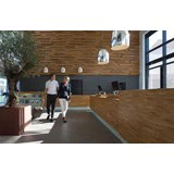 To personer går med bagage i en moderne hotellobby med trævægge, pendellamper, store vinduer og et pottetræ.