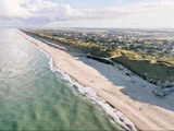 Luftfoto af en lang sandstrand omgivet af grønne klitter og en kystby under en delvis overskyet himmel. Bølger møder blidt kysten.