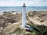 Et højt hvidt fyrtårn med en rød top står på sandede klitter nær havet, omgivet af snoede stier og græsklædt terræn under en delvist overskyet himmel.