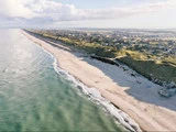 Luftfoto af en lang sandstrand omgivet af blide bølger på den ene side og græsklædte klitter med huse på den anden, under en delvis overskyet himmel.