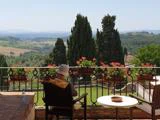 En person med solhat sidder på en terrasse med blomster og udsigt over et naturskønt landskab med bølgende bakker og træer under en klar himmel.