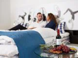Et par sidder på en seng med front mod hinanden og en bakke med rødvin, glas, vindruer og snacks på et glasbord i forgrunden.