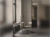 Minimalistisk arbejdsplads med metalskrivebord, justerbar lampe, stol på hjul og pyntegenstande ved et stort vindue i et neutralt rum.