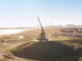 En stor trebuchet af træ står på en græsklædt bakke nær kysten med huse og et industriområde i baggrunden under en klar himmel.